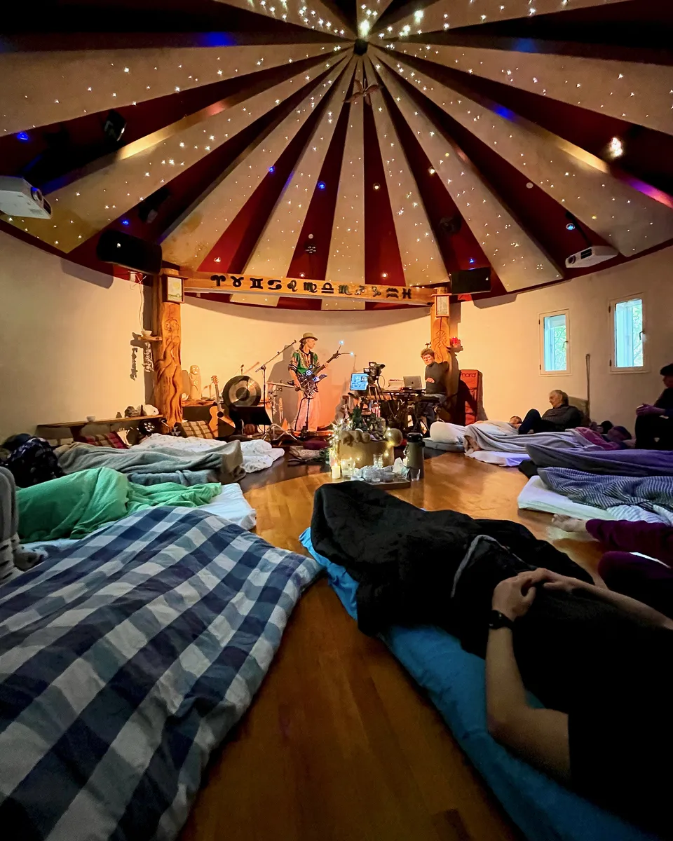 Ayahuasca ceremony in the Roundhall: participants lie on mattresses under blankets while the musicians play by the altar beneath the striped starry sky in the ceiling