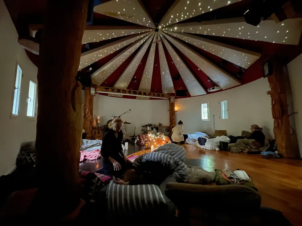 Ayahuasca-ceremoni i Rundsalen for kvinder: en guide knæler ved siden af en kvinde, der ligger under et tæppe, mens lys brænder ved alteret og stjernehimlen lyser i loftet