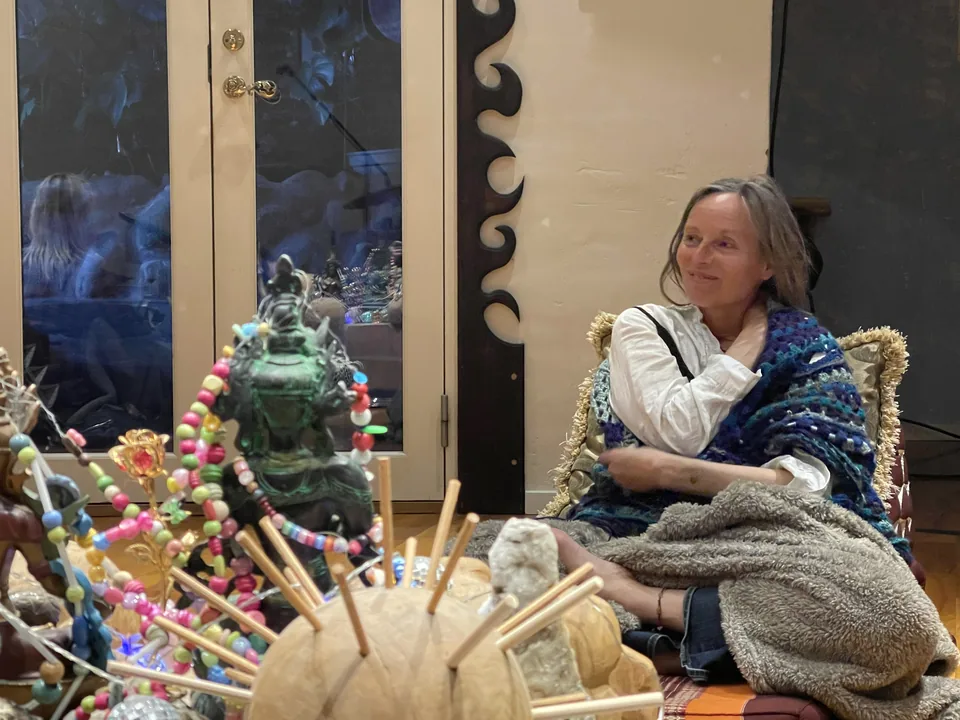 En kvinde sidder svøbt i tæpper ved alteret i Rundsalen efter ayahuasca-ceremonien og smiler stille, med rasler og små figurer foran sig