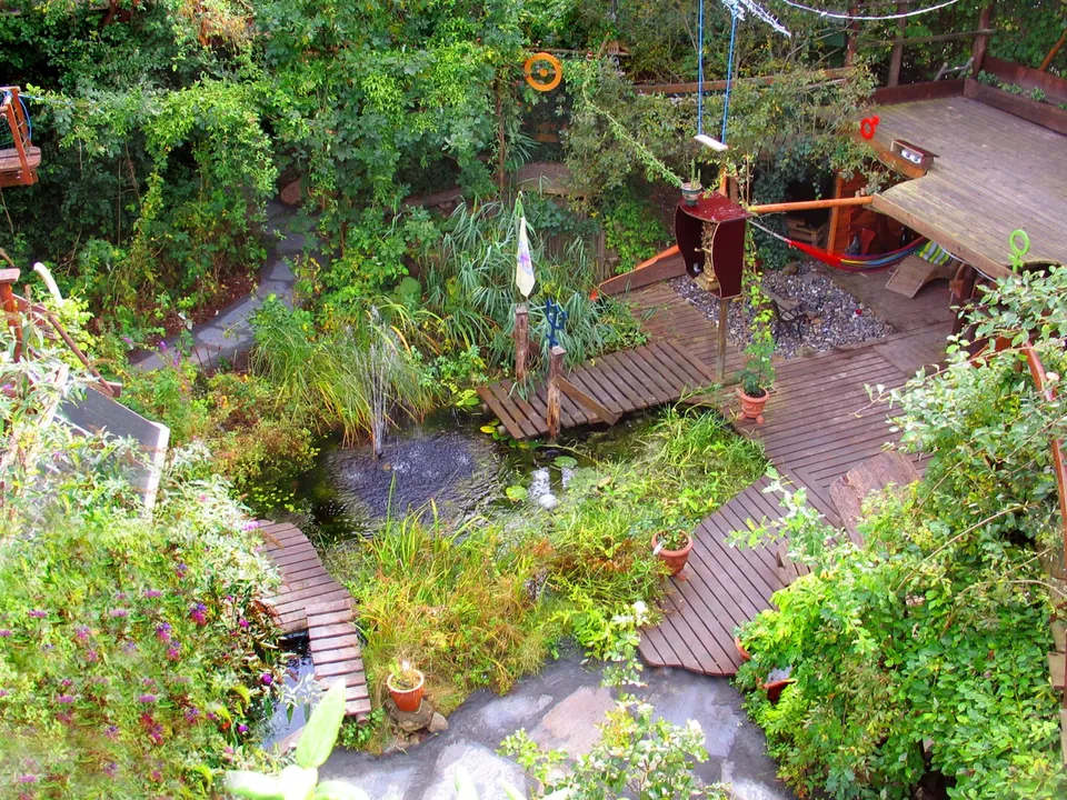 The Magic Garden set oppefra: en lille sø med springvand, omgivet af træbroer, stier af skifer, tætte planter og en terrasse med en hængekøje
