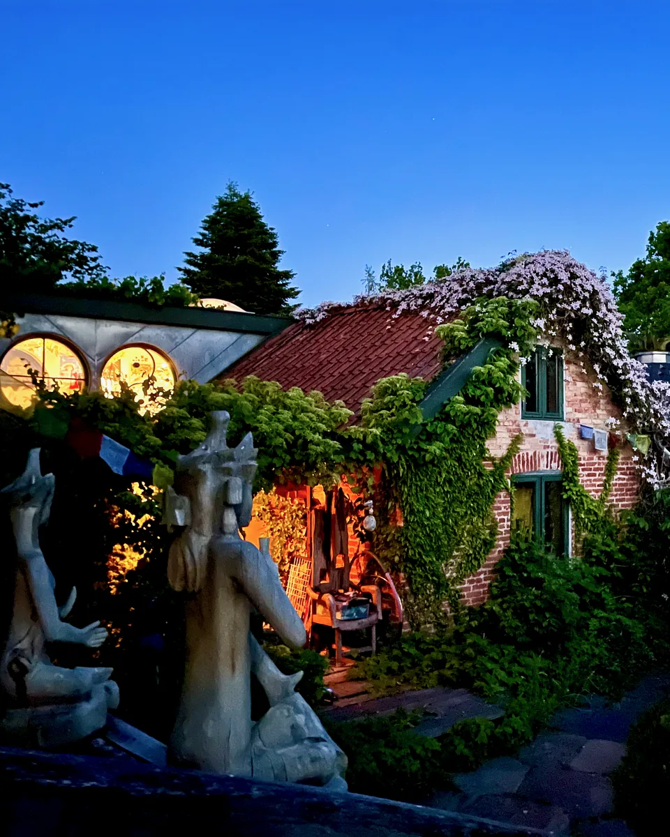 Huset og Rundsalen i Fredensborg, hvor vi holder ayahuasca retreat for kvinder i Danmark, i skumringen under en dybblå himmel, med lys i de runde vinduer, blåregn på taget og stenskulpturer forrest