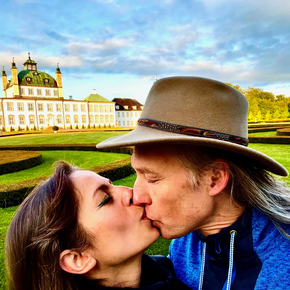 Jacob og Ecaterina Moth kysser i slotshaven ved Fredensborg Slot i 2022, 22 år efter deres bryllup. Han bærer hat, og slottet ses bag dem.