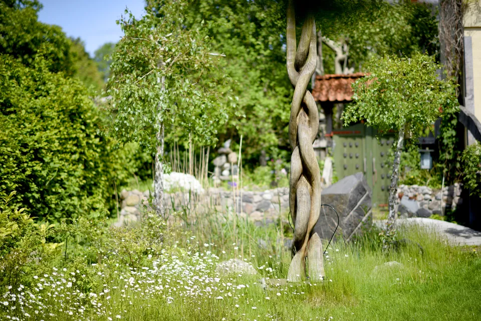En snoet træsøjle i haven mellem markblomster, med havens port i baggrunden