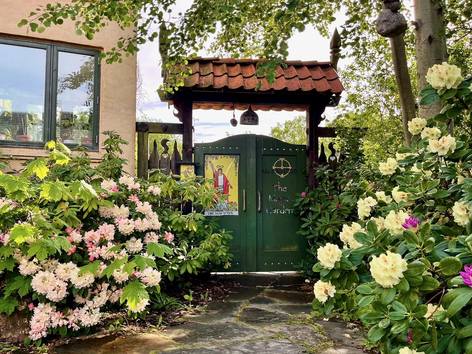Havens grønne port med tarotkortet Magikeren, omgivet af blomstrende rhododendron en forårsdag