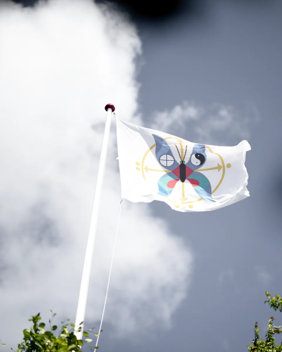 Et hvidt flag med The Magic Gardens sommerfugl vajer mod blå himmel og hvide skyer over haven